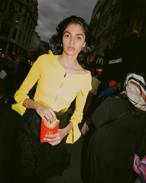 A young woman in a yellow top eats French fries in a city street scene, with a McDonald's restaurant sign visible in the background.