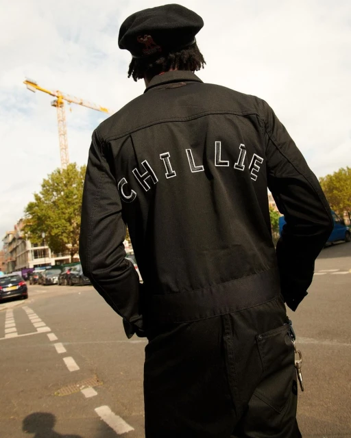 Adult poses on city street wearing black jumpsuit and beret, showcasing back design near yellow construction crane.
