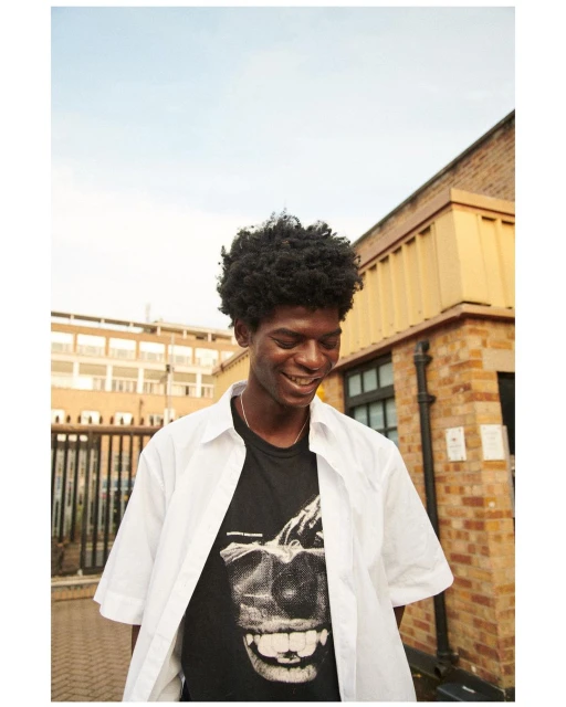 Portrait of a smiling Black man with an Afro wearing a graphic tee and open white shirt, standing in front of brick buildings and metal gates.