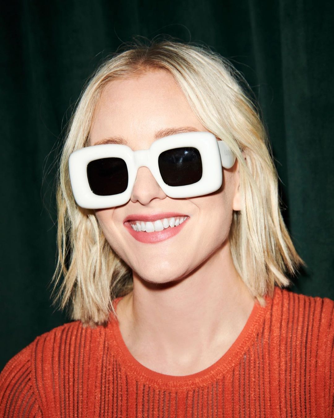 Close-up portrait of a smiling woman with short blonde hair wearing an orange top and large, white, square-framed sunglasses.