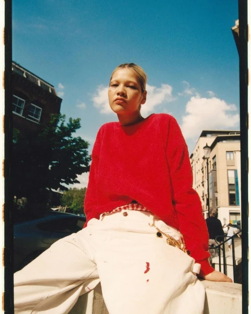 Young woman in casual red sweater and white pants poses outside, bathed in sunlight against a blue sky with buildings in the background.