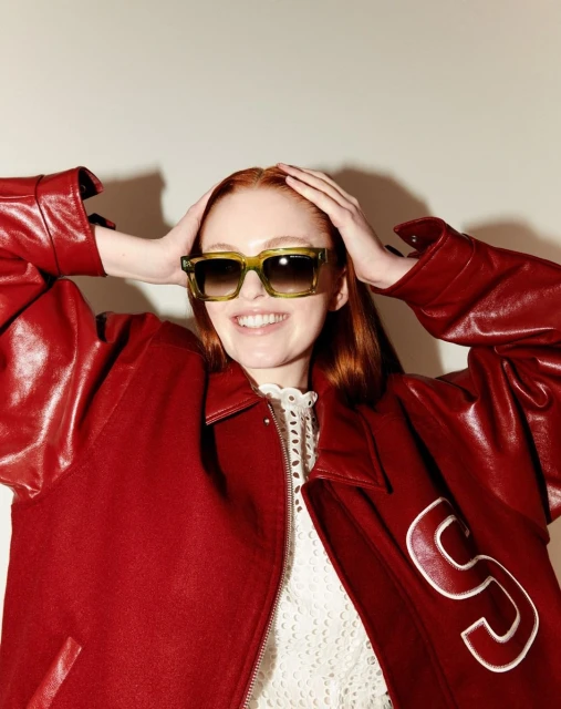Smiling woman with auburn hair wearing sunglasses and a red leather jacket indoors.  Close-up shot of model in fashion setting.