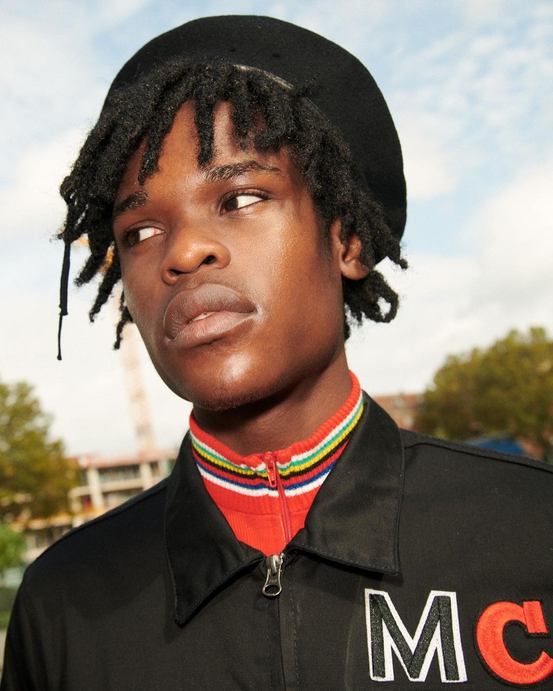 Portrait of stylish Black male model in beret, wearing a black jacket with embroidered letters MC, photographed outdoors against a building.