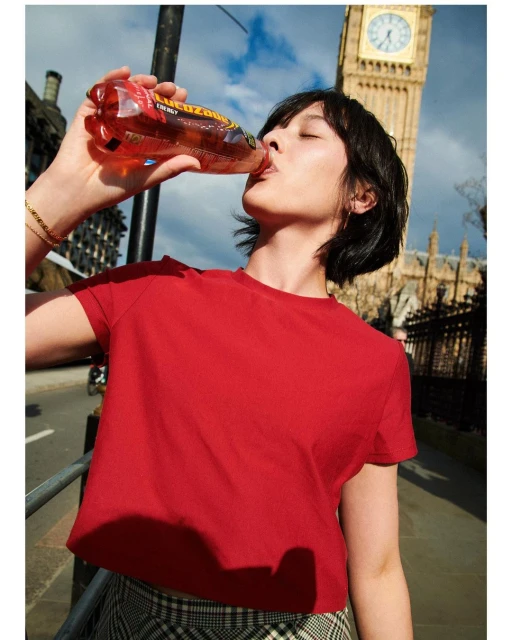 Woman drinks a Lucozade beverage outdoors in front of Big Ben. She wears a red top and a plaid skirt in the city.