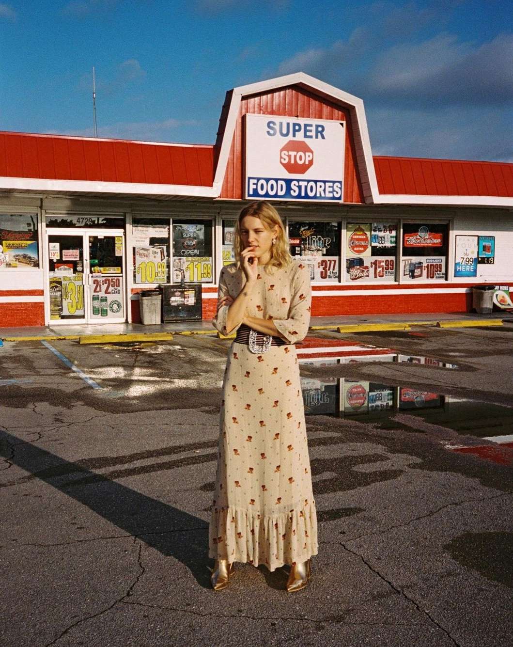 A blonde woman in a long floral dress stands in front of a Super Stop Food Store on a sunny day, posing with a contemplative expression.