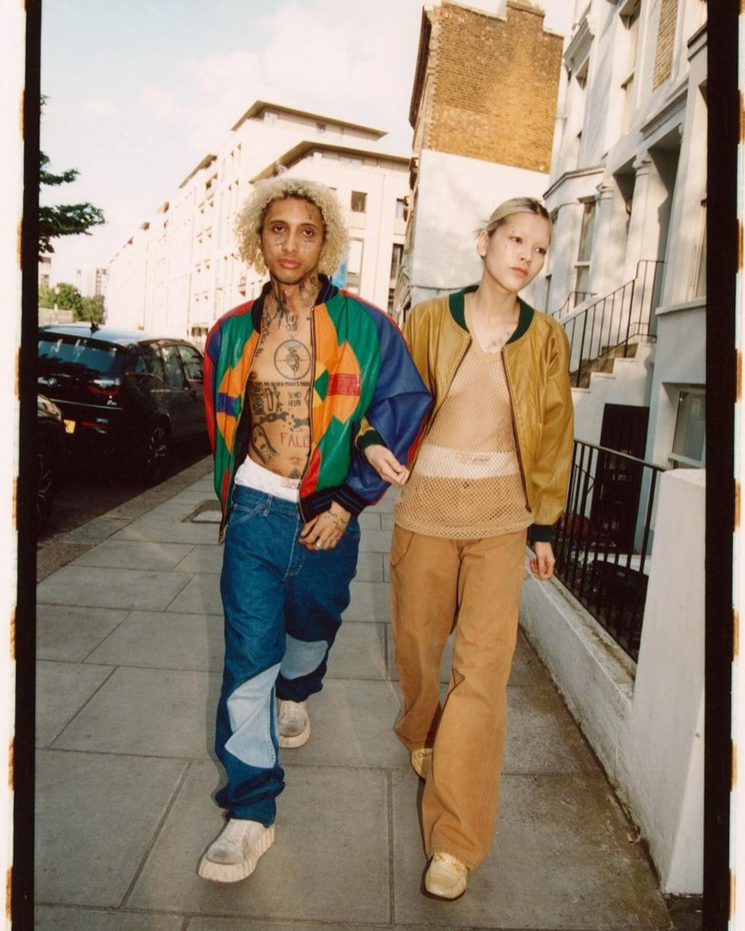 Two fashion models stroll an urban street. One wears a colorful jacket and jeans, the other a tan jacket with matching pants.