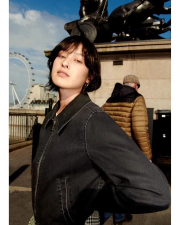 A young woman in a denim jacket stands in an urban setting, with a bronze statue and the London Eye in the background, bathed in daylight.