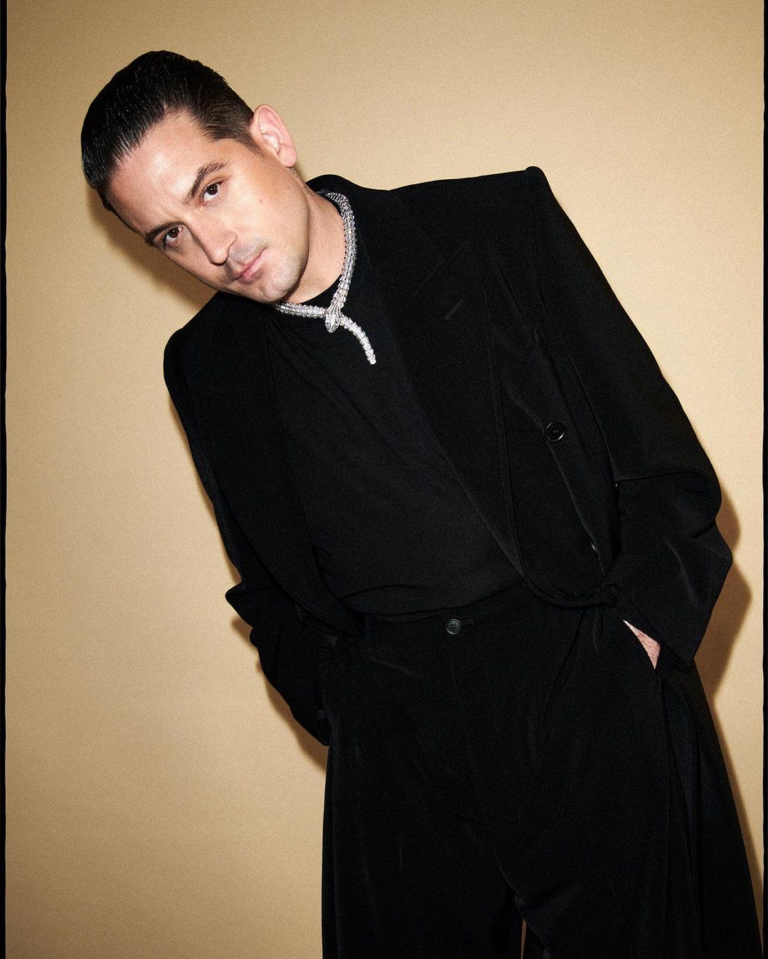 Portrait of a handsome adult male in a black velvet suit and white gold necklace, posing against a tan studio backdrop.