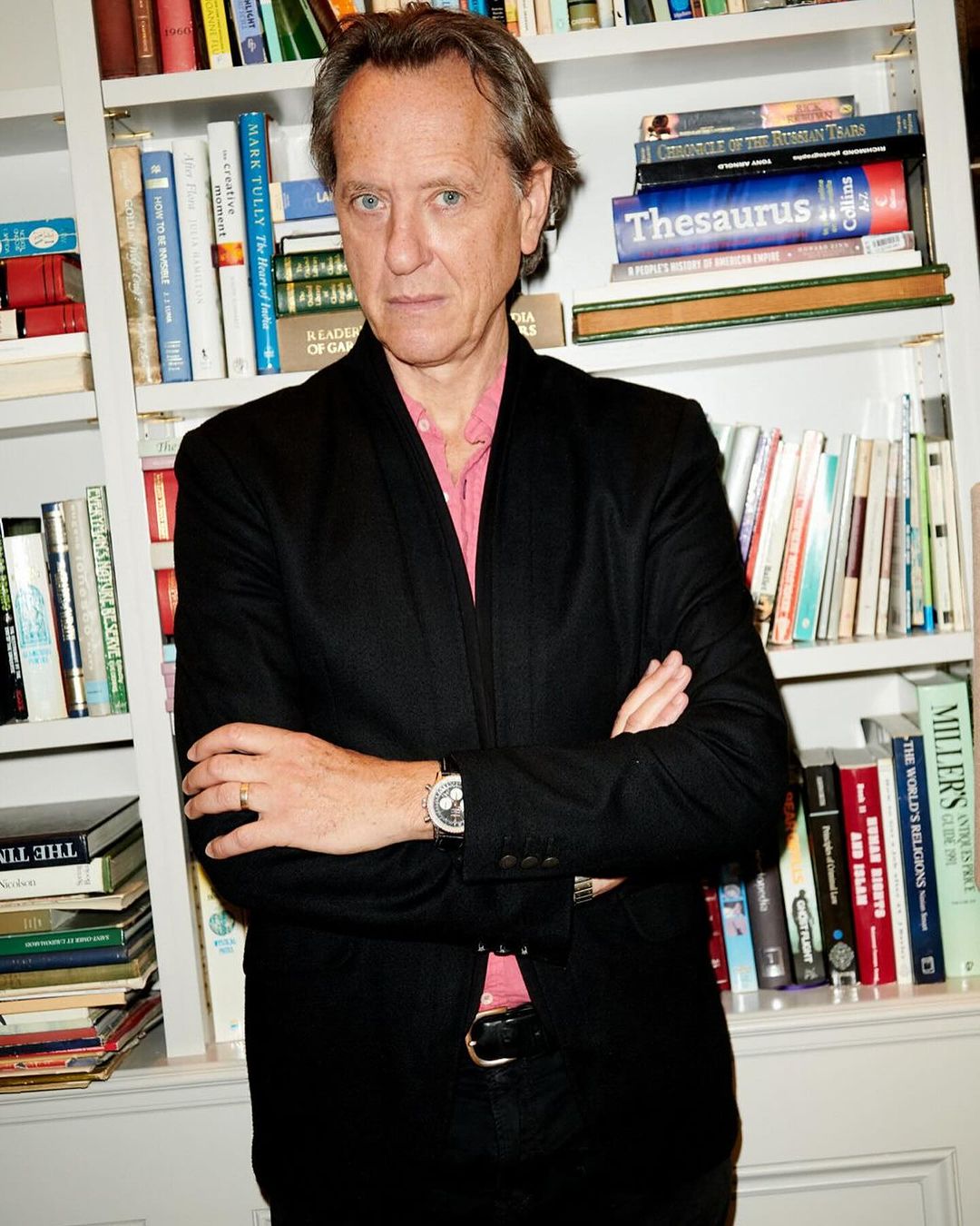 Mature adult man in a black suit and pink shirt poses indoors in front of a bookshelf filled with books, wearing a watch and ring.