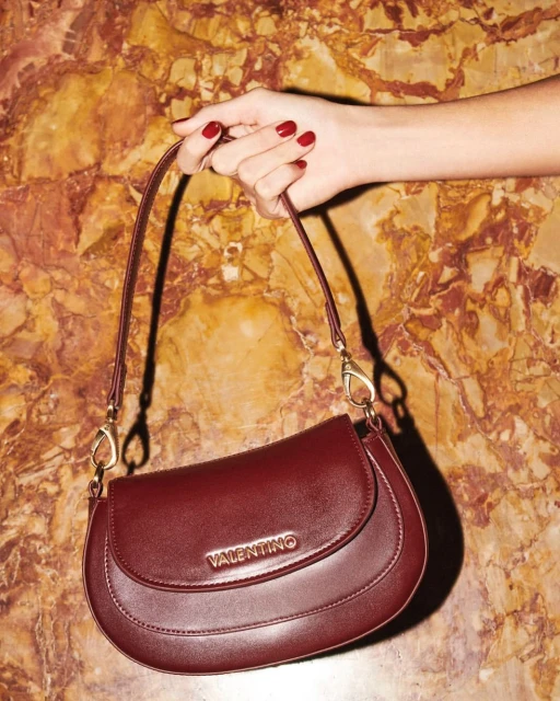 Burgundy Valentino handbag with gold logo and hardware held by a fair-skinned hand with red nail polish against a marble background.