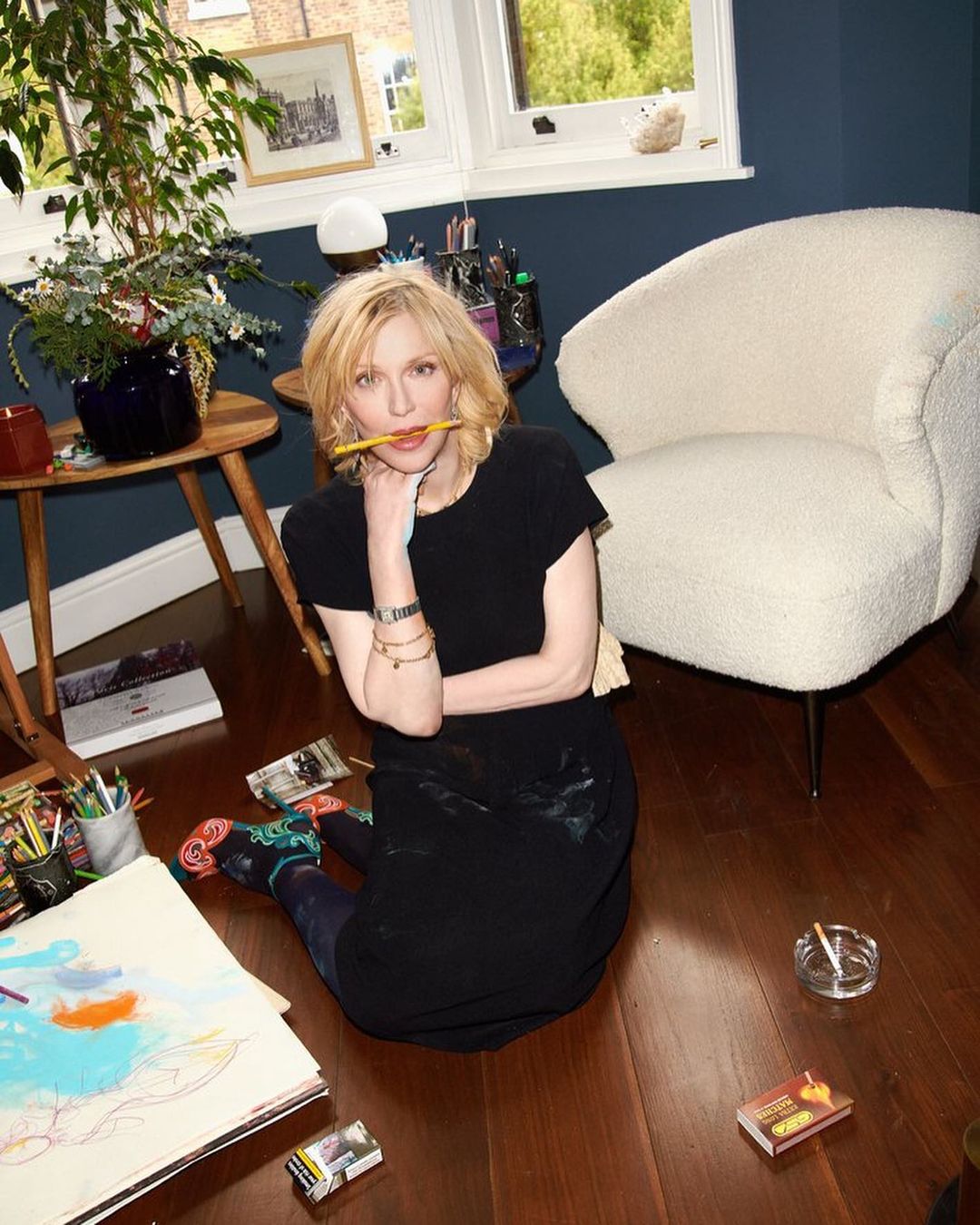 Indoor portrait of a blonde adult female sitting on a wooden floor with a pencil in her mouth, surrounded by art supplies.