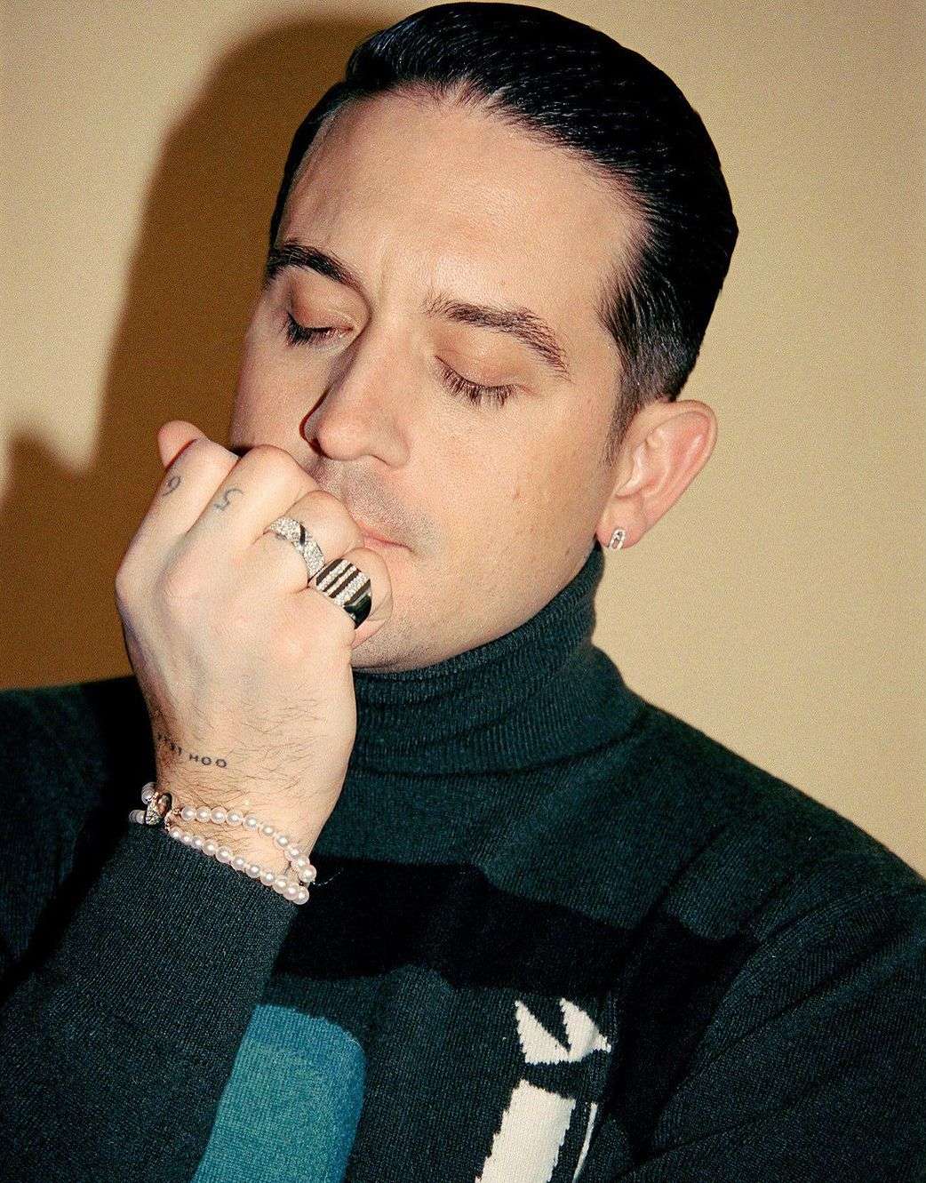 Portrait of a handsome adult male wearing a turtleneck sweater, rings, and pearl bracelets, with his fingers to his lips.
