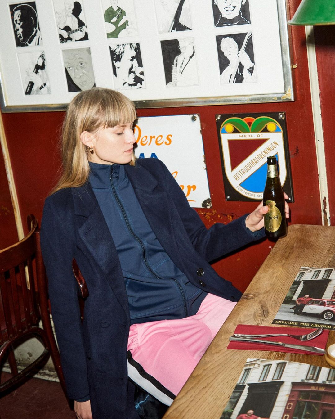 Indoor shot of a blonde woman holding a beer, wearing a blue jacket and pink pants, sitting at a wooden table with magazine on top.