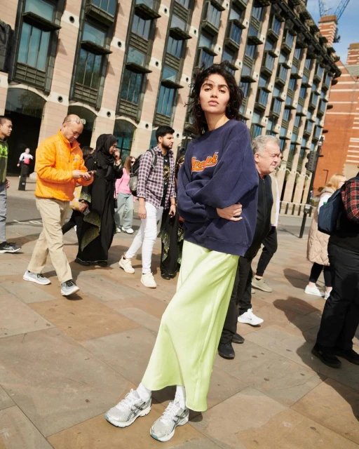 A stylish young woman in a blue sweatshirt and green skirt poses on a city street with a crowd of people and a brick building behind her.