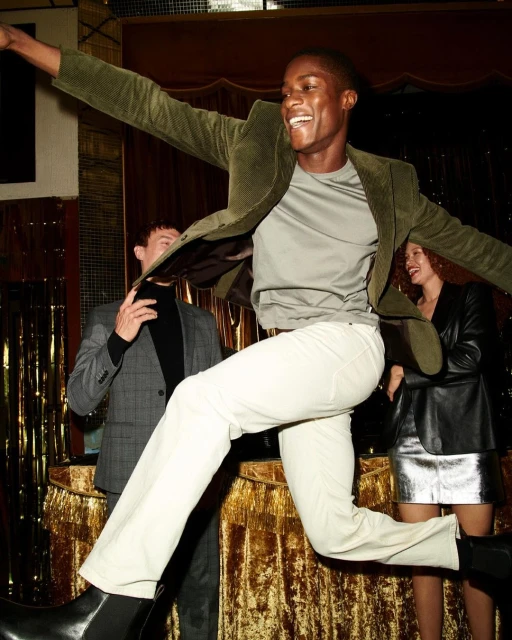 A joyful Black man in a green blazer leaps in the air indoors, while two other people look on, in a nightlife or celebration setting.