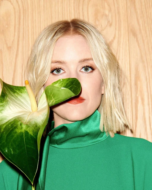 Close-up of a blonde woman in a green dress posing with a green Anthurium flower against a light wood background indoors.
