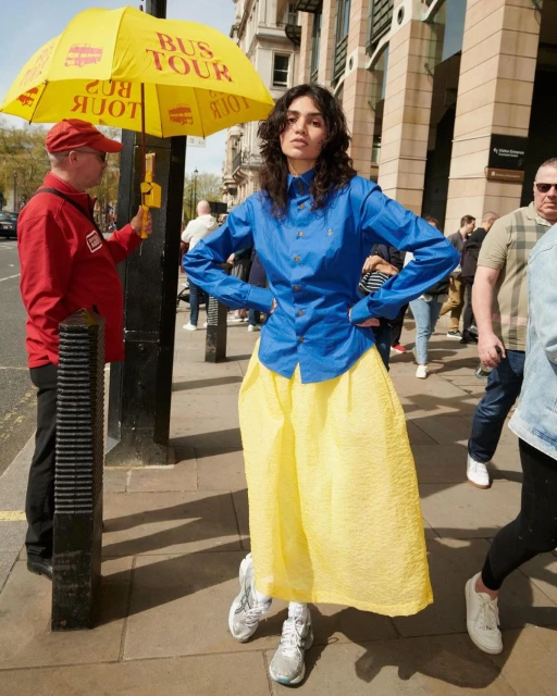 Fashionable woman in a blue shirt and yellow skirt poses on a city street next to a bus tour guide under a yellow umbrella.