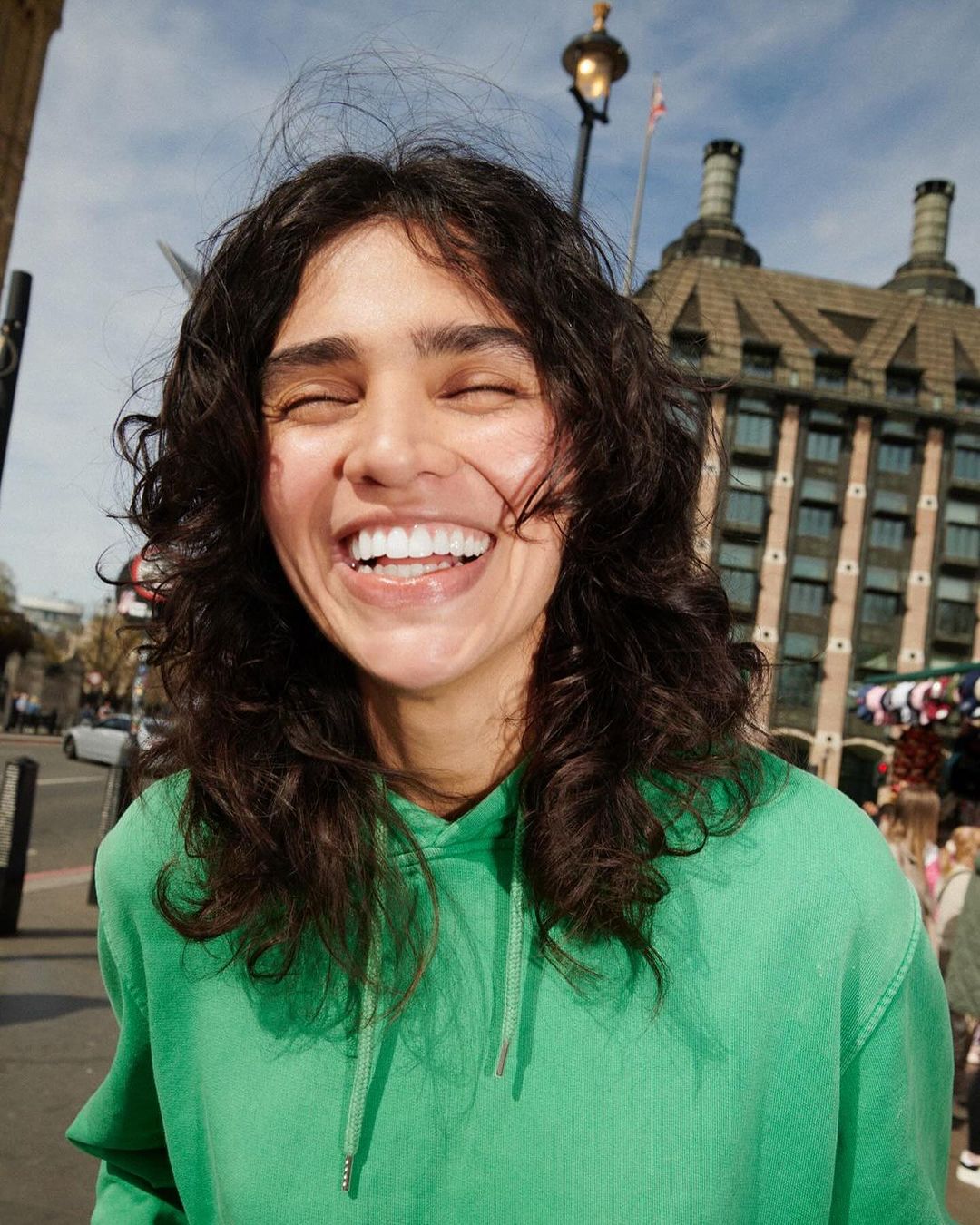 Close-up of smiling woman with curly hair wearing green hoodie outdoors with buildings and street visible in the background.