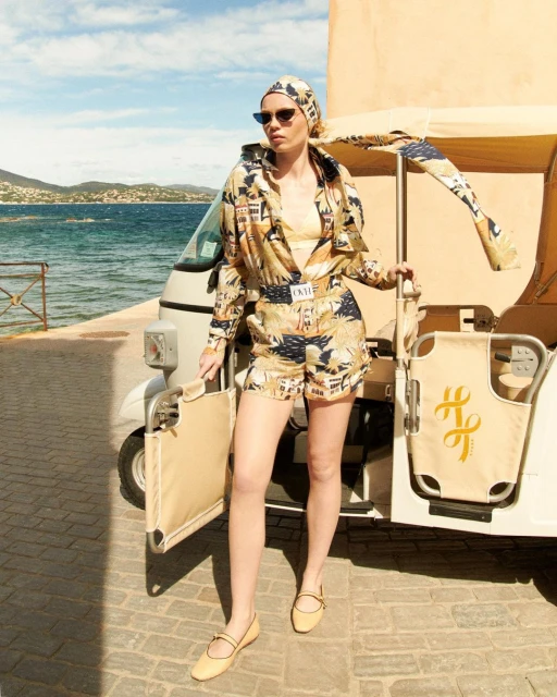 A fair-skinned adult female model poses in a patterned two-piece outfit near a three-wheeled vehicle by the sea on a sunny day.