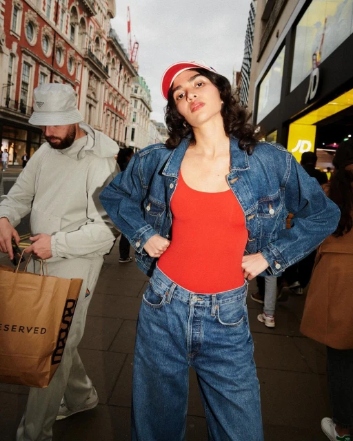 A woman wearing a red top and jeans poses in an urban setting while a man walks by carrying a shopping bag.