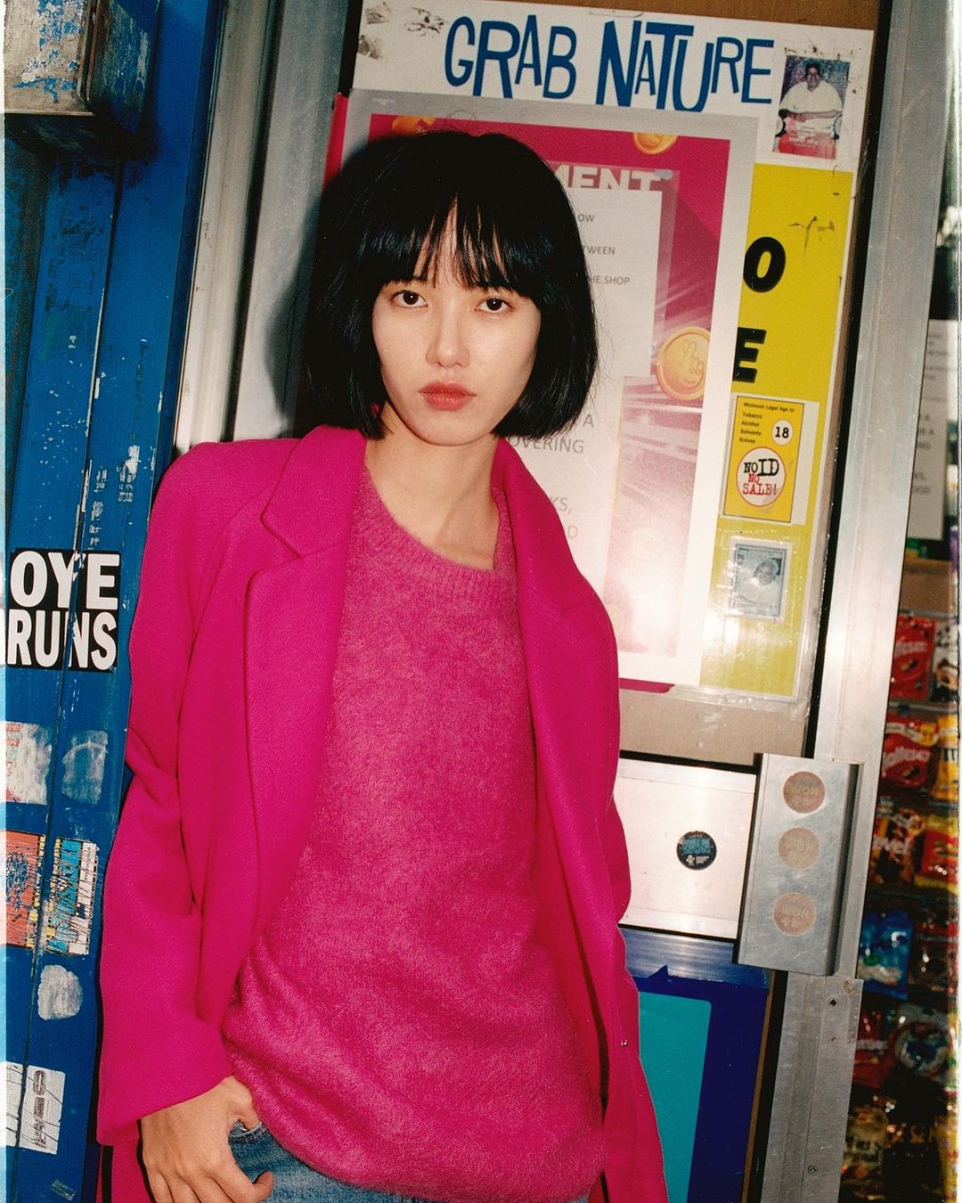 An adult woman stands next to a newsstand wearing a pink sweater and blazer, set in an urban environment with bright, natural lighting.