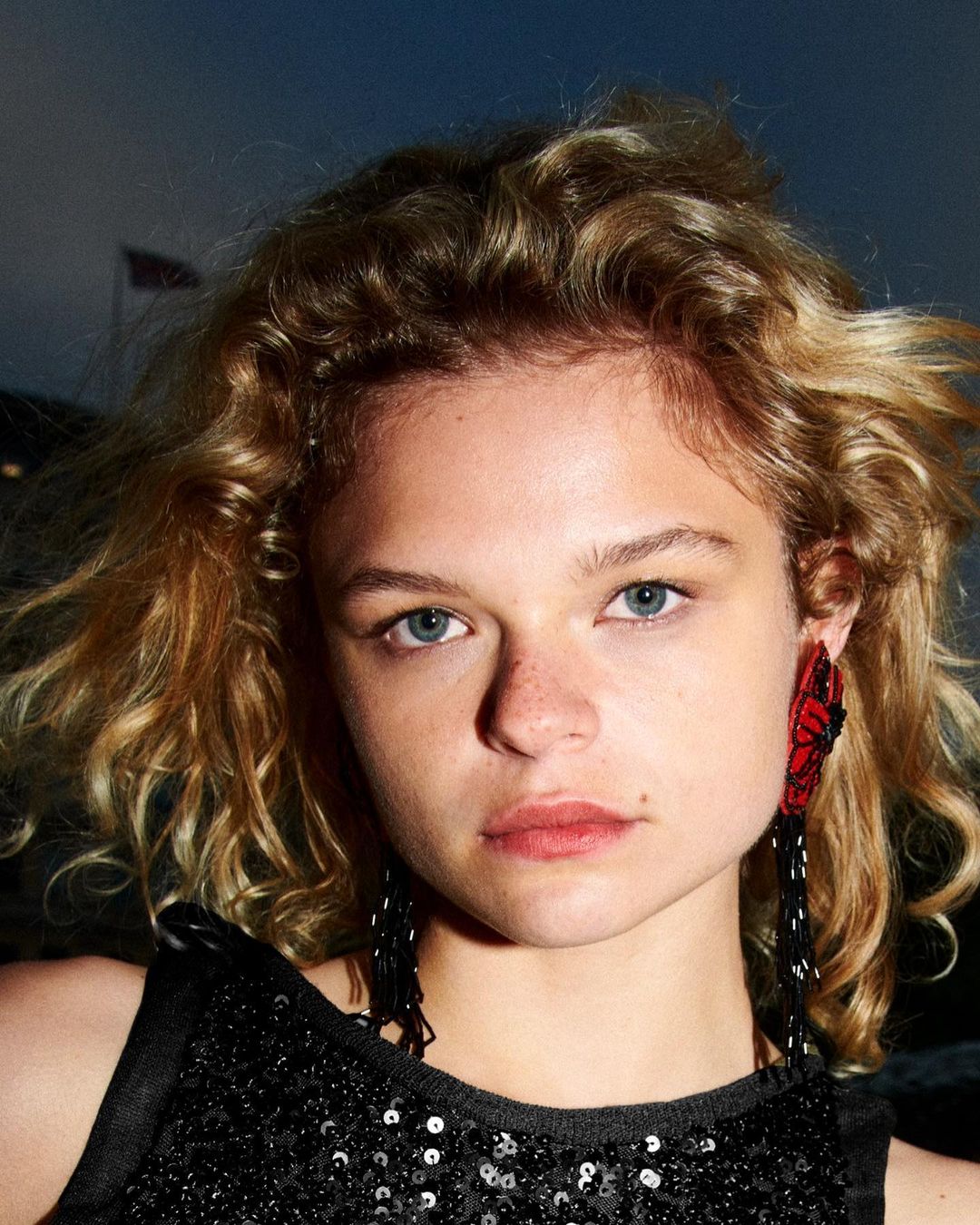 Close-up portrait of a young woman with curly blonde hair wearing a black sequined top and red floral earrings with long beaded strands.