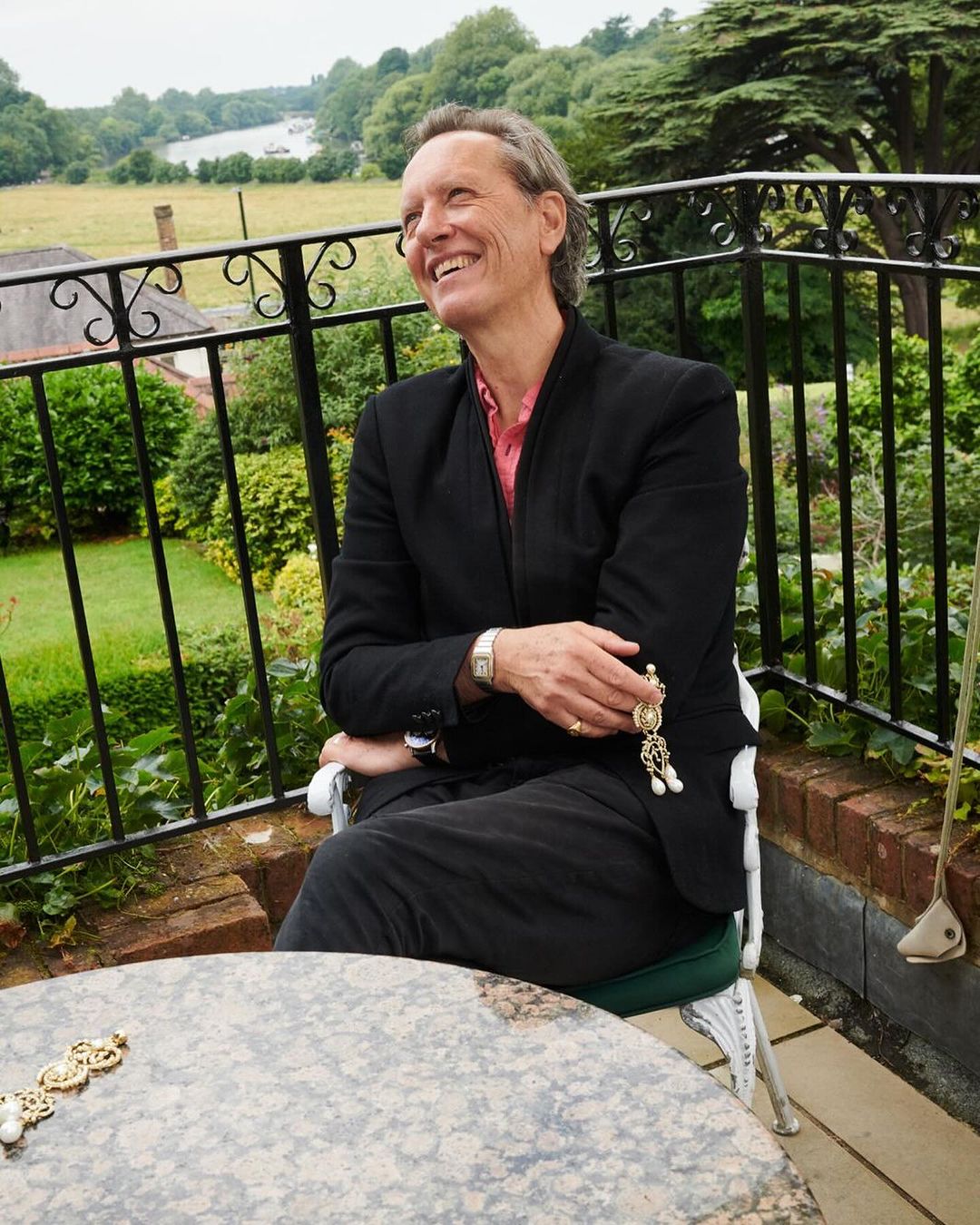 Smiling man in a blazer sits on a balcony with a green landscape. Jewelry visible. Outdoor shot with greenery and sky backdrop.