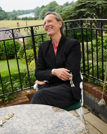 Middle-aged man in a black blazer and pink shirt sitting on a balcony overlooking a green landscape, photographed by Andrew Woffinden.