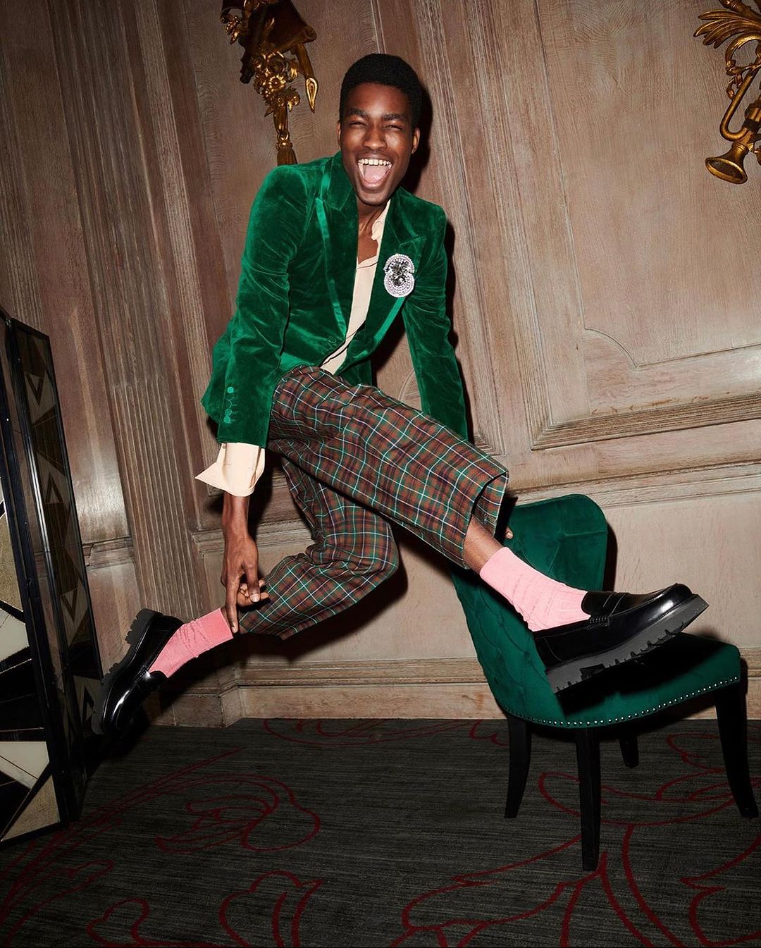 Joyful African American man in a green velvet jacket leaps from a chair in an ornate room, wearing plaid pants, pink socks, and black loafers.