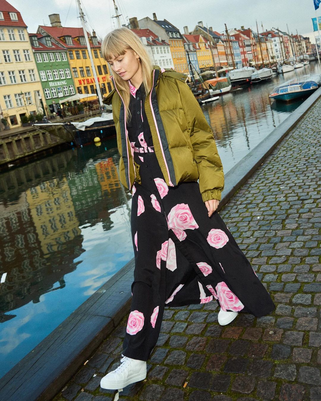 Blonde woman in an olive puffer jacket and floral dress standing on a cobblestone walkway with canal and colorful buildings in the background.