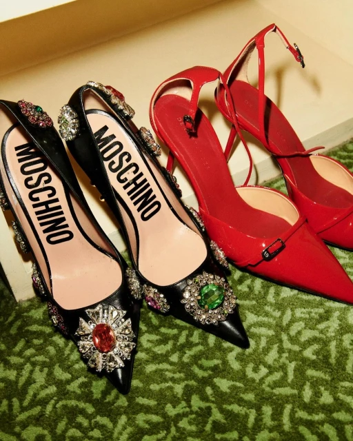 Elevated view of two pairs of high heels, one black with jewels and Moschino logo, the other red patent leather, on a green surface.