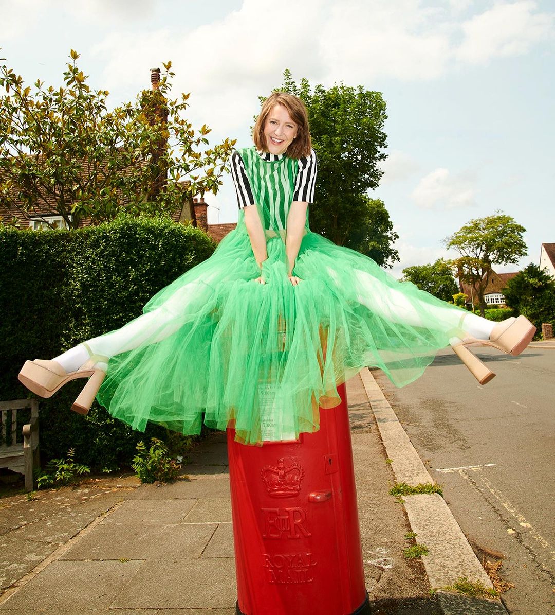 Young woman in green tulle skirt and striped top sits in a straddle pose atop a red British postbox on a sunny suburban street.
