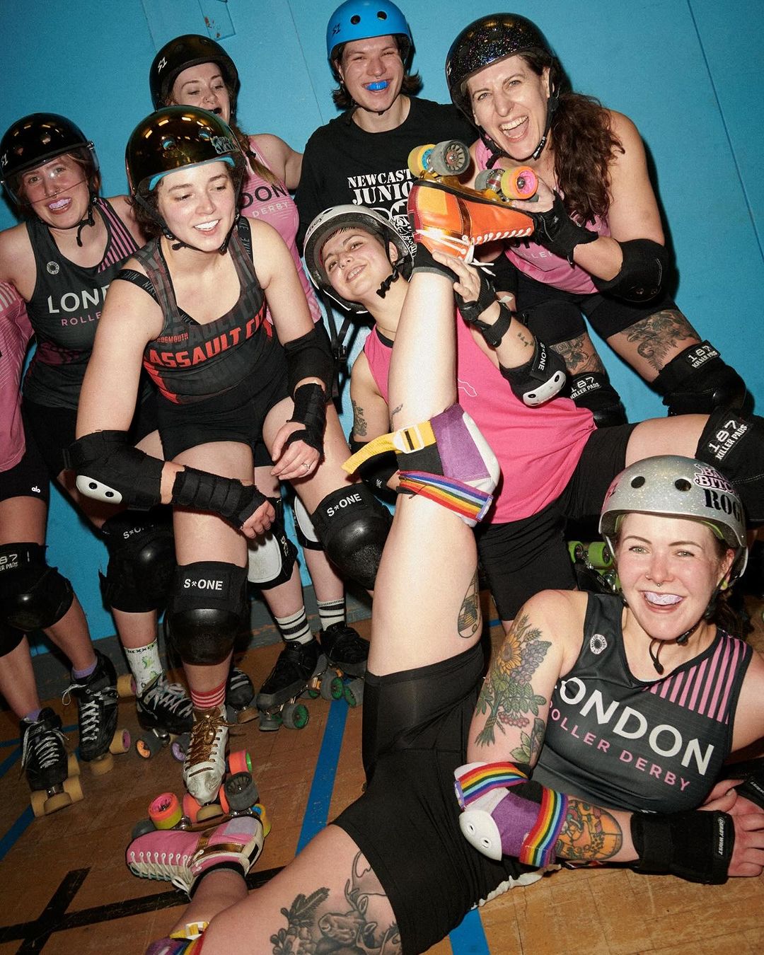 Group of seven roller derby athletes wearing helmets, pads, and skates smiling and posing indoors against a blue wall.