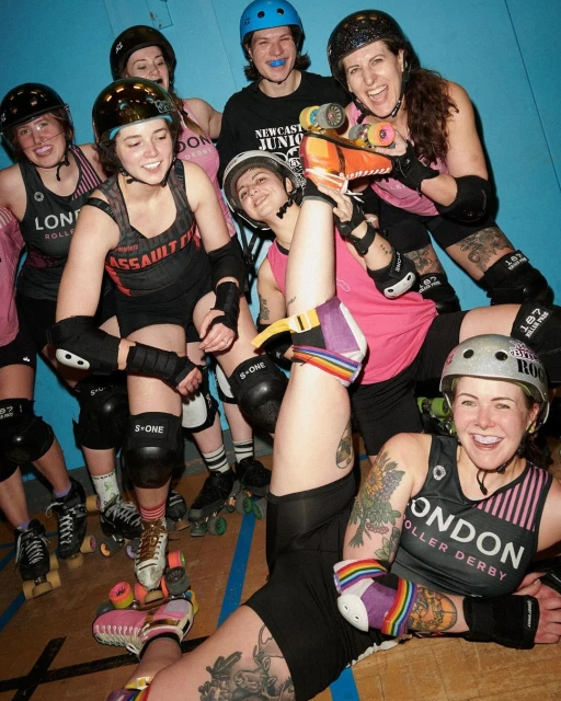 Group of seven roller derby athletes wearing helmets, pads, and skates smiling and posing indoors against a blue wall.