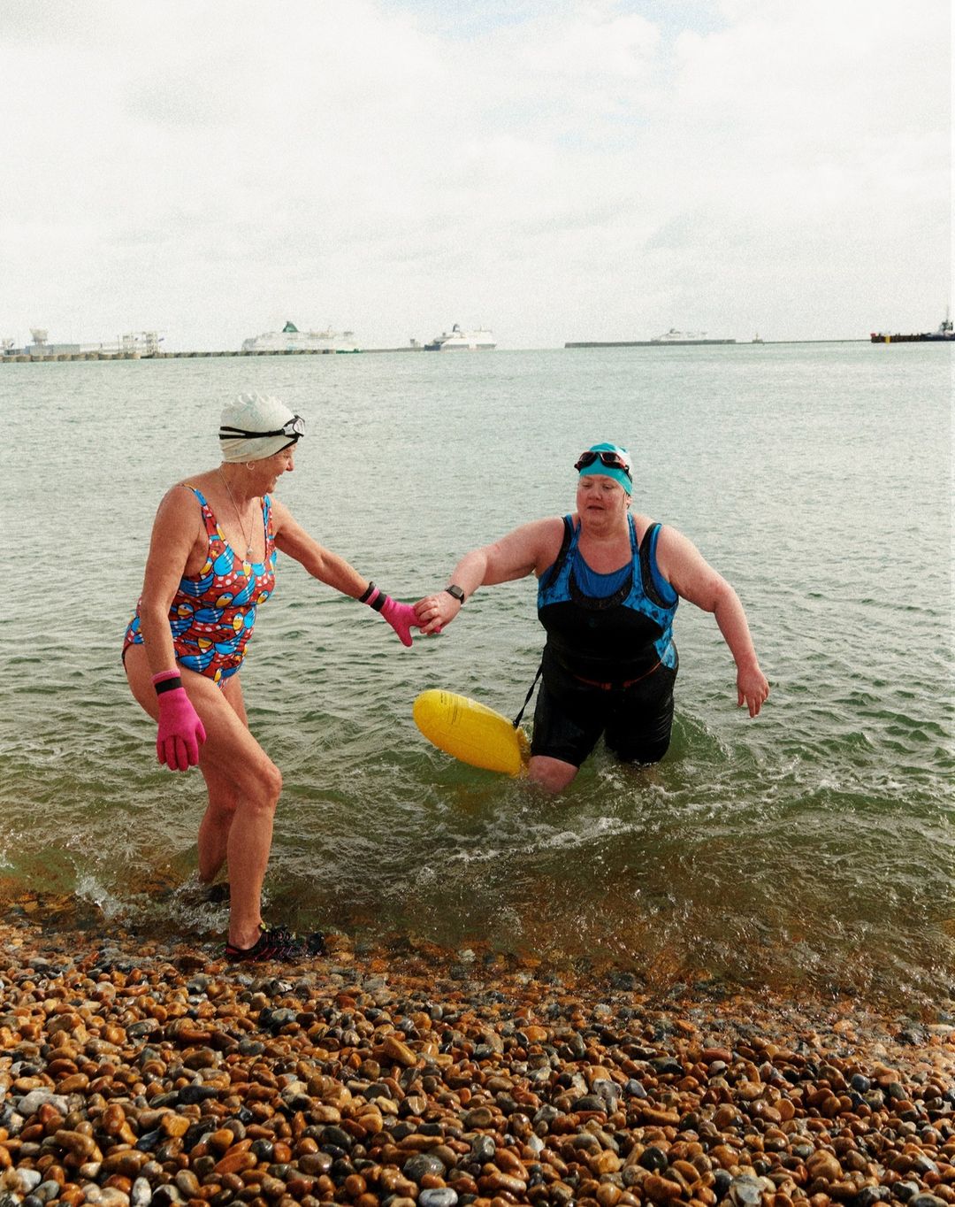 Two women in swimwear enter the ocean, one holding the other's hand, with a yellow safety buoy in clear daylight by a pebble beach.