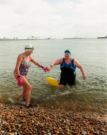 Two women in swimwear enter the ocean, one holding the other's hand, with a yellow safety buoy in clear daylight by a pebble beach.