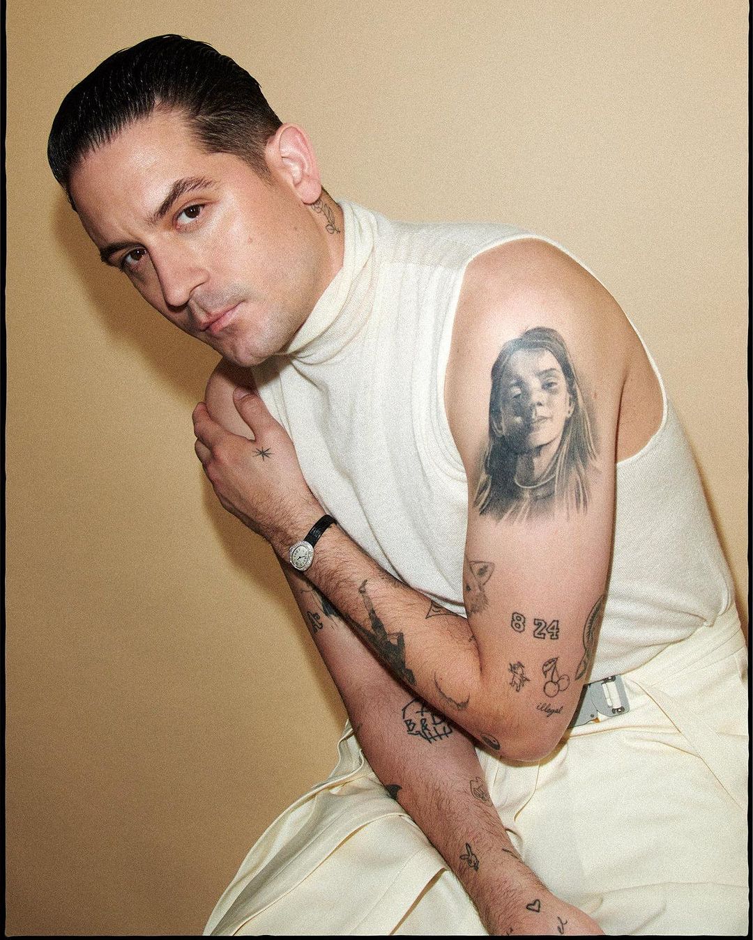 Portrait of a man with an undercut, wearing a white sweater and off-white pants, showcasing tattoos on his arms against a tan background.