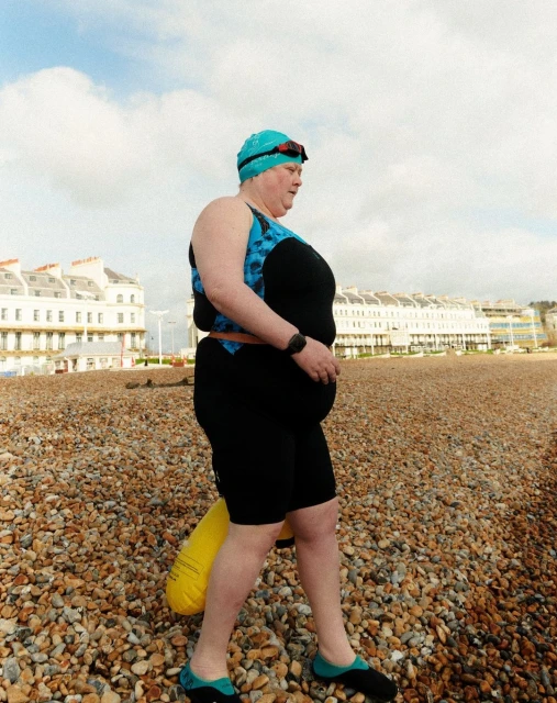 Female swimmer in wetsuit walking on pebble beach, wearing swim cap and goggles, carrying a yellow float, with buildings in background.