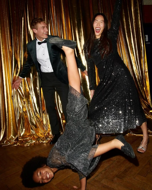 Three people in formalwear pose on a wooden floor with a gold background; one person is doing a handstand.