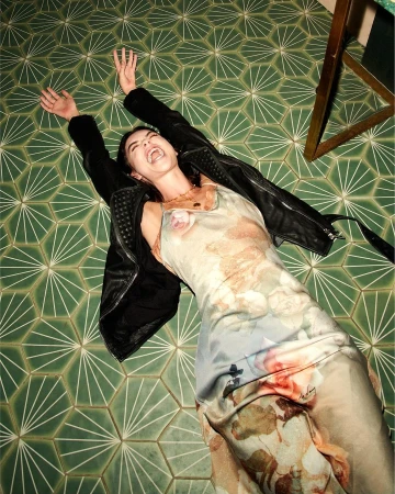 Woman in floral silk slip dress and black leather jacket lying flat on green geometric tiled floor with hands raised.