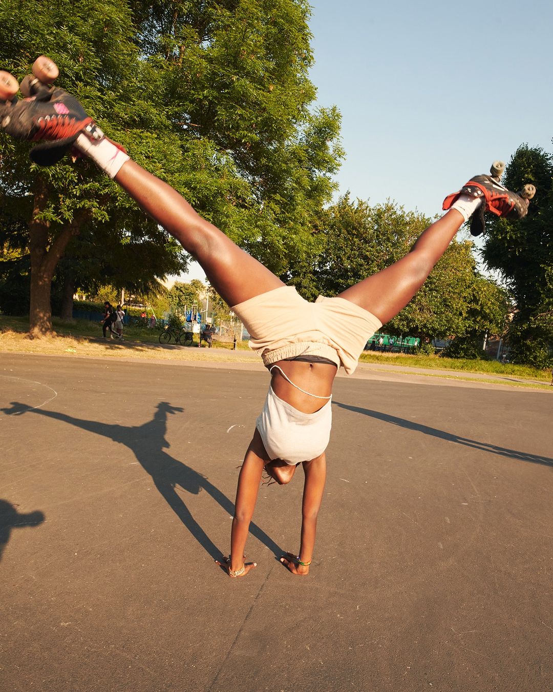 A dark-skinned adult woman is doing a handstand on an asphalt surface wearing roller skates in a bright outdoor setting in a park.