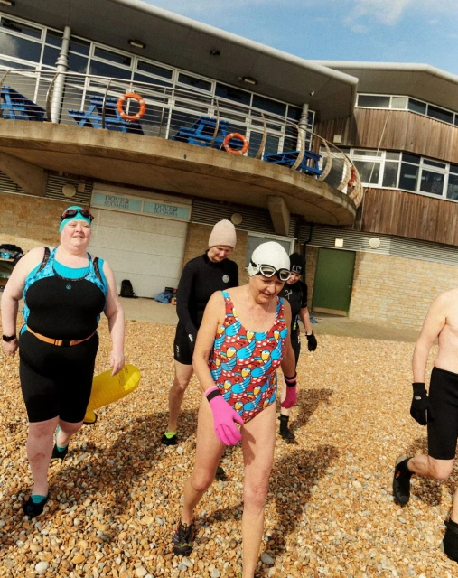 A group of older adult open water swimmers in swimsuits and goggles walk on a pebble beach near a low building, under a bright sky.