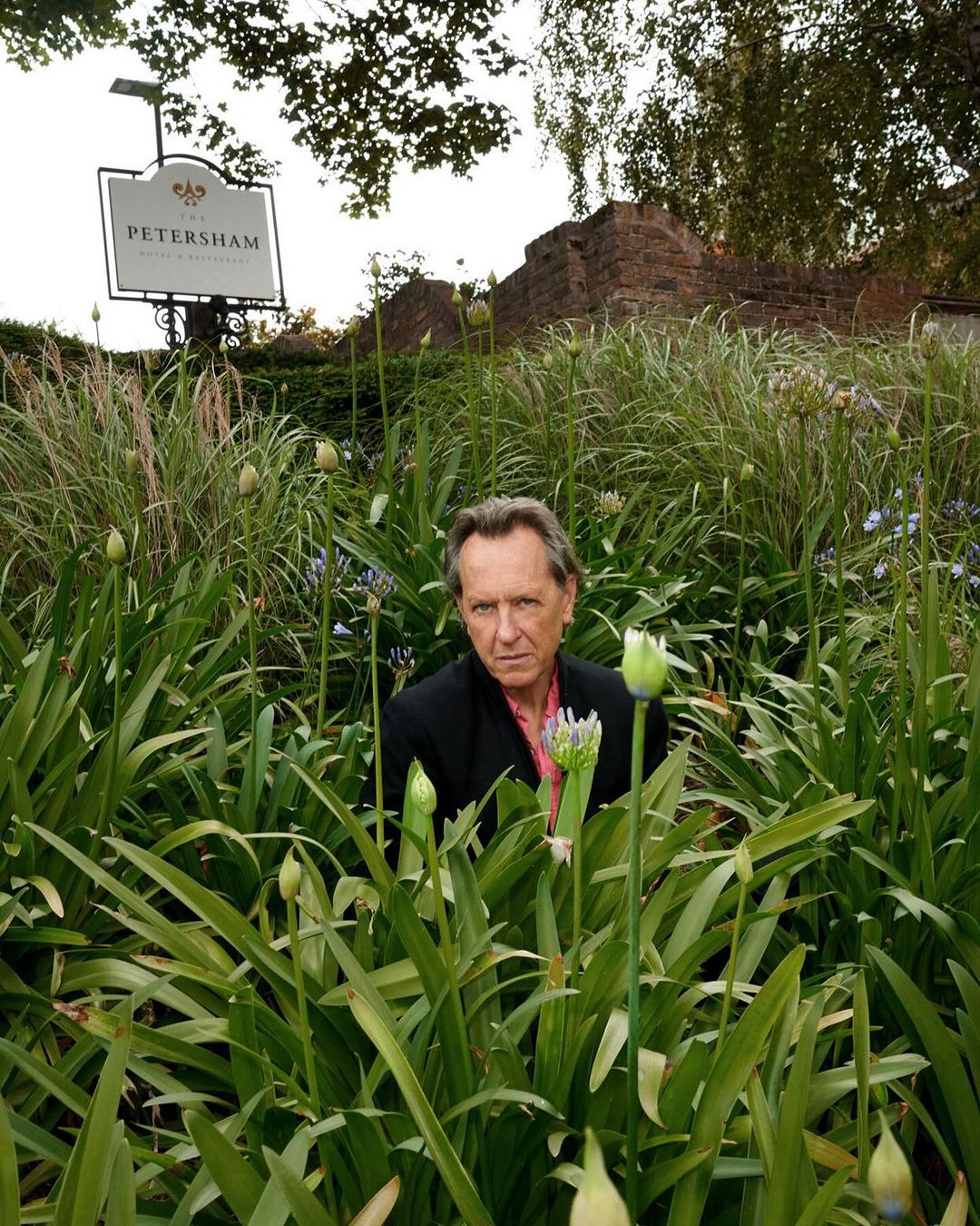 Portrait of an adult male standing in a garden with blooming plants and a sign in the background on a cloudy day.