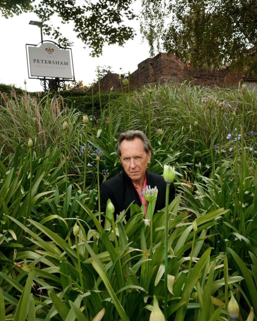 Portrait of an adult male standing in a garden with blooming plants and a sign in the background on a cloudy day.