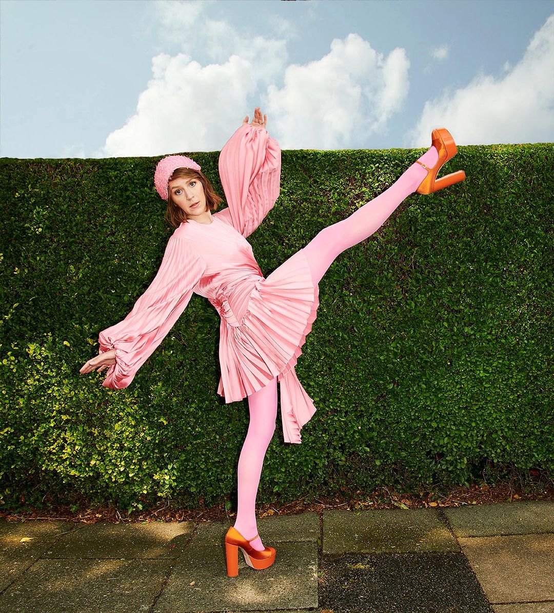 A young adult female in a pink dress and orange platform shoes strikes a dynamic pose against a green hedge under a partly cloudy sky.