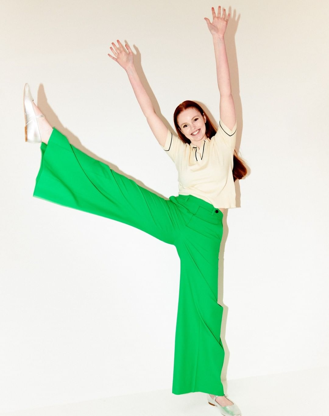 A vibrant, full-length portrait of a woman in green pants and a cream-colored top, smiling with raised arms and legs, set against a clean white backdrop.