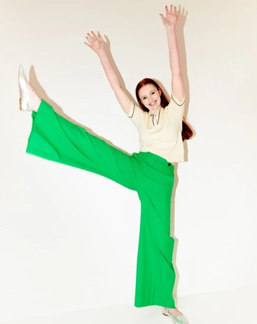 A vibrant, full-length portrait of a woman in green pants and a cream-colored top, smiling with raised arms and legs, set against a clean white backdrop.
