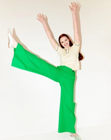 A vibrant, full-length portrait of a woman in green pants and a cream-colored top, smiling with raised arms and legs, set against a clean white backdrop.