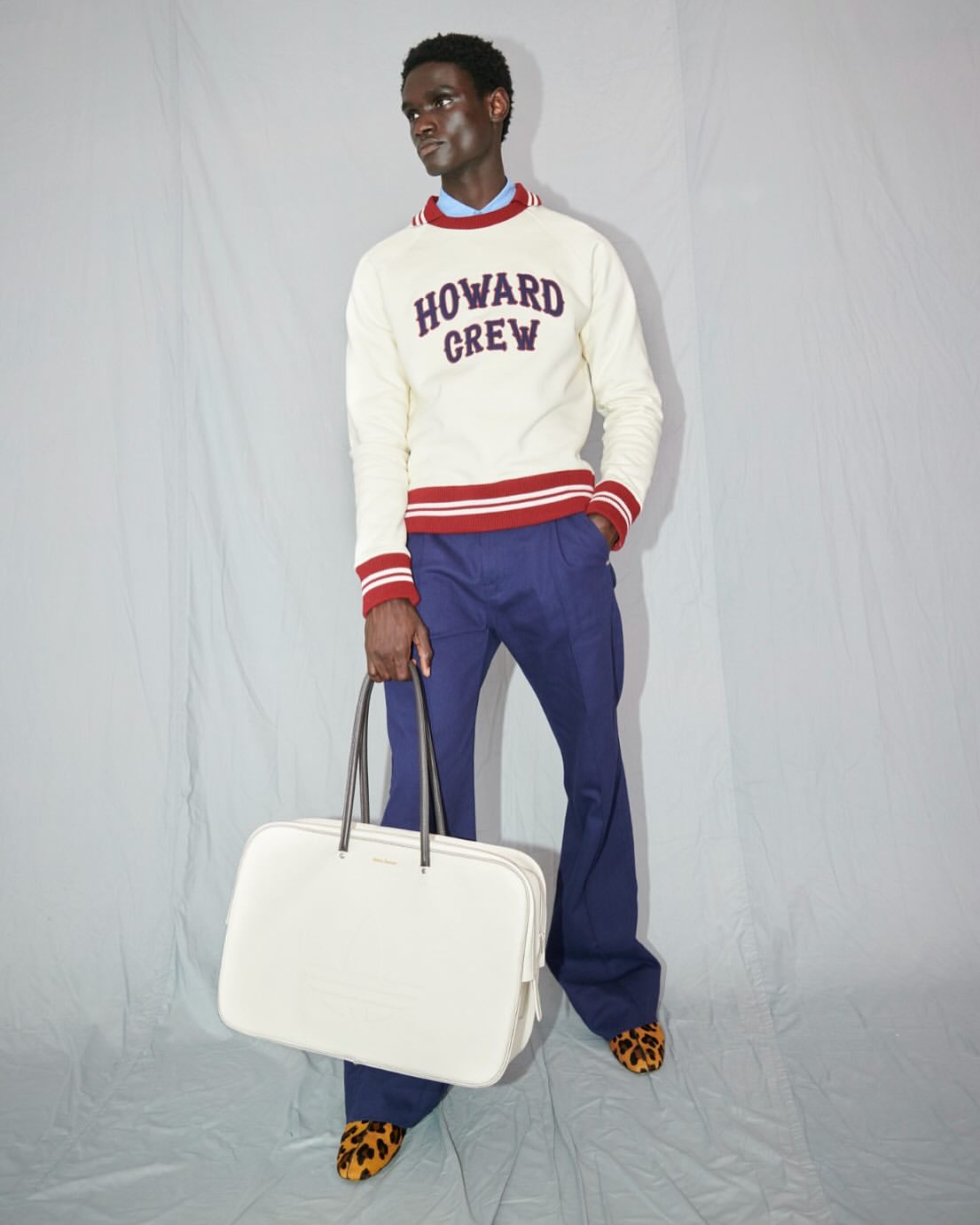 Male fashion model posing in a cream-colored sweater and blue pants, holding a large white bag in a photo studio.