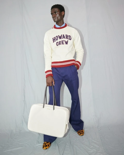 Male fashion model posing in a cream-colored sweater and blue pants, holding a large white bag in a photo studio.